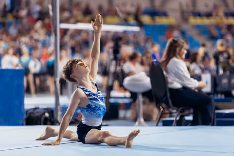 Young gymnast performs a split pose on the floor mat, arm raised gracefully upward, face lifted with focused expression.