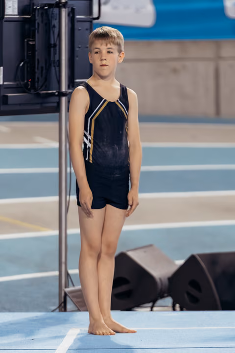Young male gymnast stands barefoot on blue floor, poised and focused, wearing black and gold leotard before his routine.