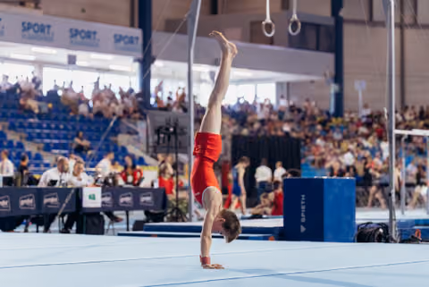 Male gymnast in red leotard holds a perfect handstand on the floor exercise mat before a packed arena crowd.