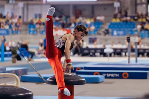 Young male gymnast performs a split on the pommel horse, leaning forward with intense concentration at an indoor competition.
