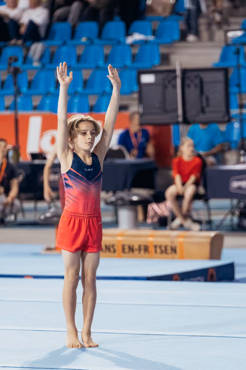 Young gymnast stands barefoot on the mat, arms raised high in a confident salute, wearing a red and navy leotard.