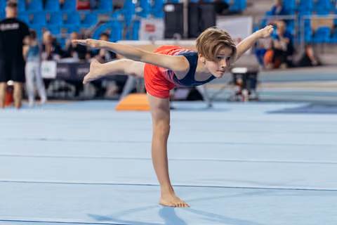 Young gymnast holds a focused arabesque balance on one leg, arms extended, on a blue competition floor.