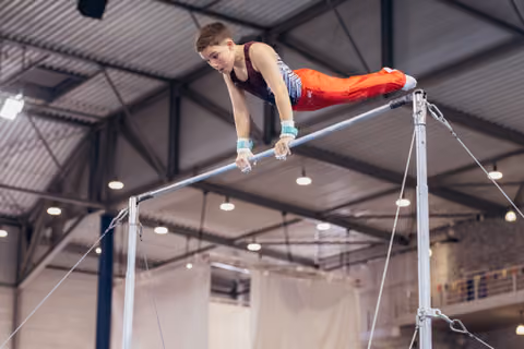 Young male gymnast performs on horizontal bar, body parallel, intense focus, inside a large indoor gymnasium.