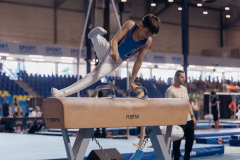 Young male gymnast performs on pommel horse, leaning forward with intense focus during an indoor competition.