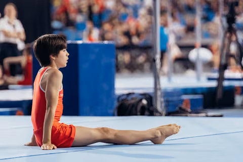 Young gymnast sits in a straddle position on the blue floor mat, composed and focused, during a competition.