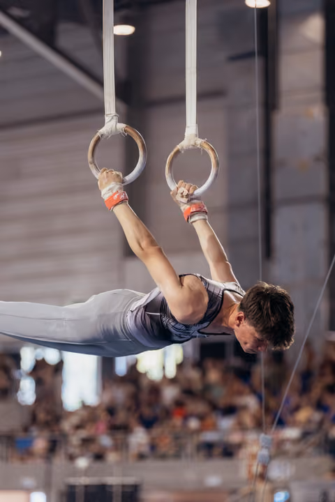 Male gymnast performs an iron cross on still rings, body horizontal, showing remarkable strength and concentration at a gymnastics meet.