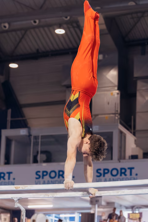 Young male gymnast performs a precise handstand on parallel bars, body fully extended upward in red competition leotard.