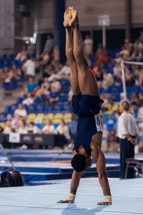 Male gymnast performs a perfect handstand on floor exercise, body fully extended, competing before a live audience.