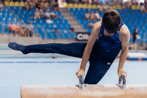 Young male gymnast performs scissors move on pommel horse, head down in deep concentration during competition.
