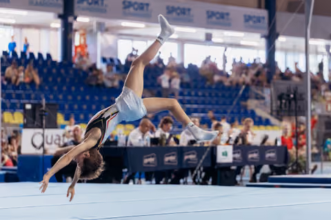 Young gymnast mid-backflip on floor exercise, body fully extended, competing before a packed arena crowd.