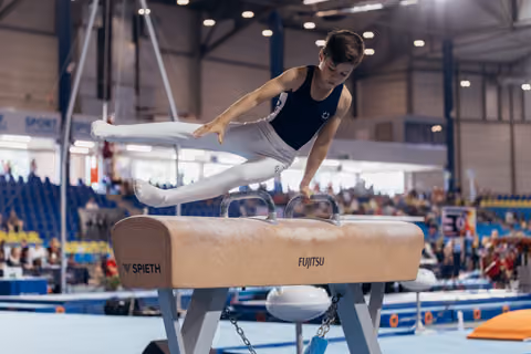 Young male gymnast performs a precise pommel horse routine, legs split wide, intense focus, in a packed competition arena.