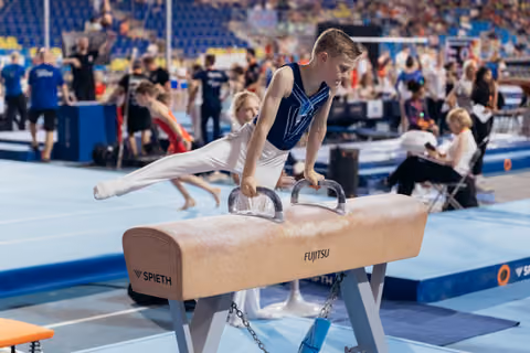 Young male gymnast performs a focused pommel horse routine, legs extended, at a busy indoor gymnastics meet.