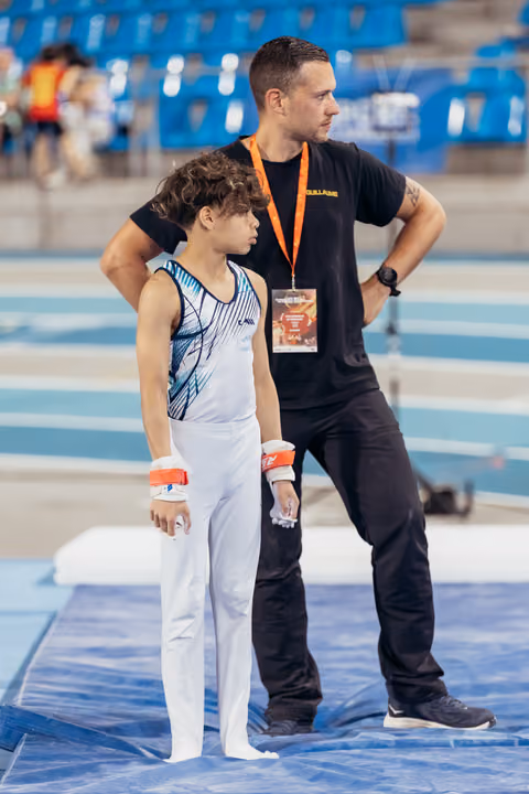 Young gymnast stands focused beside coach in black, both observing the floor exercise area at an indoor sports arena.