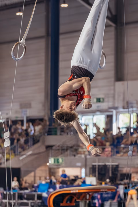 Male gymnast performs an inverted hang on still rings, body fully extended, during a packed indoor gymnastics competition.