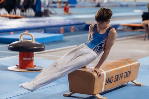 Young male gymnast sits focused on a pommel horse, concentrating during training in a blue-matted gymnasium.