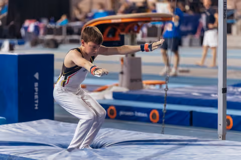 Young male gymnast lands on mat after high bar release, arms outstretched, focused expression in blue arena setting.
