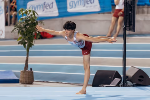 Young male gymnast holds a precise arabesque balance on the floor, arms outstretched, focused expression at a Gymfed event.