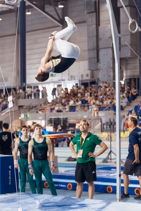 Gymnast performs an inverted flip mid-air while coach watches intently below, teammates observing in a packed competition arena.