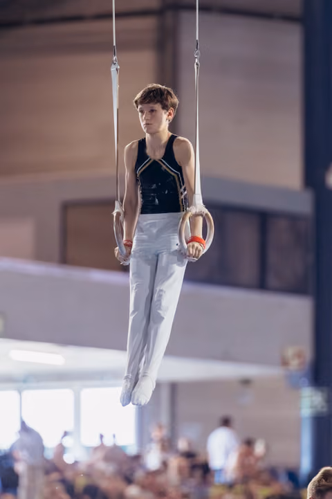 Young male gymnast holds still on rings, focused expression, wearing black leotard and white pants in a competition hall.