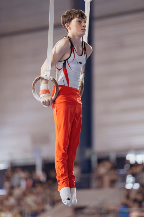 Young male gymnast performing on still rings, holding a static position with intense focus in a competition hall.
