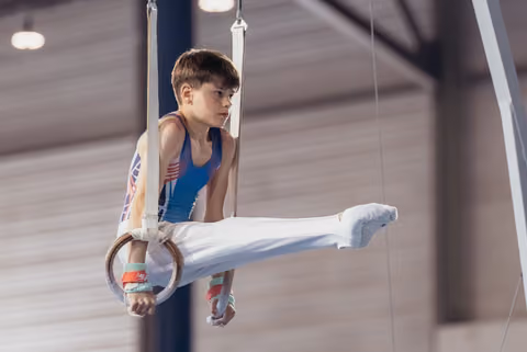 Young male gymnast performs an L-sit on still rings, wearing a blue leotard, with intense concentration on his face.