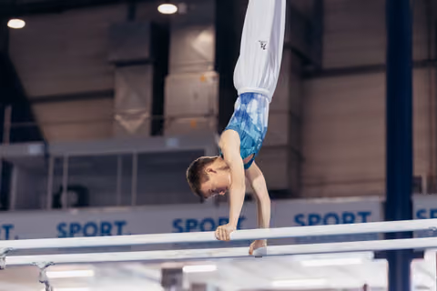 Young male gymnast performs an inverted handstand on parallel bars, showing intense focus and body control in a sports hall.