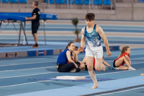 Young male gymnast runs barefoot with focused expression across blue athletics floor, teammates resting in background.