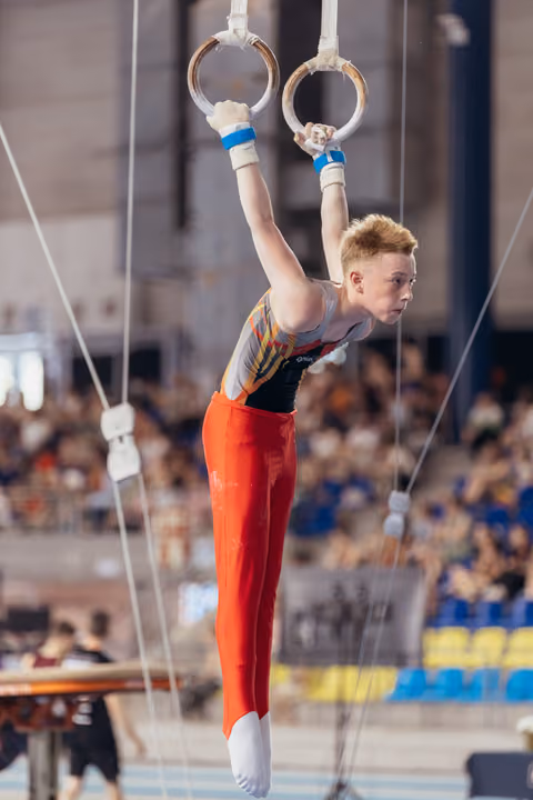 Young male gymnast grips still rings with intense focus, wearing red pants and colorful leotard in packed arena.