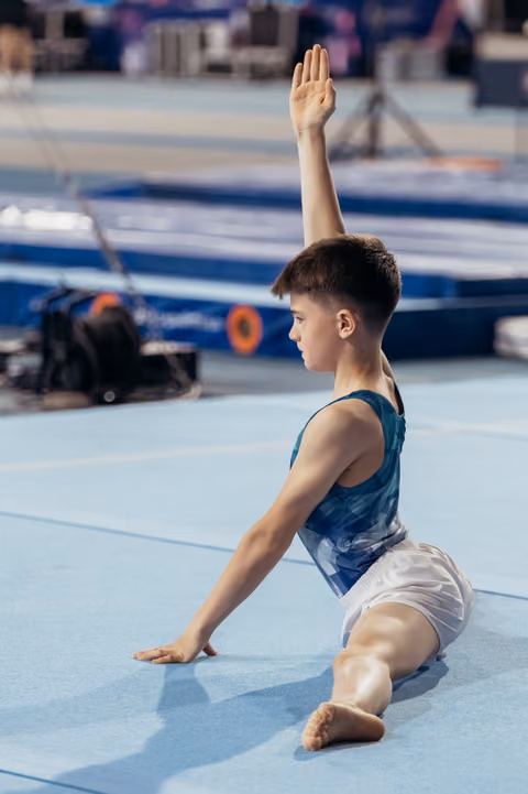 Young male gymnast sits on blue floor mat, one arm raised gracefully, focused expression during gymnastics event.