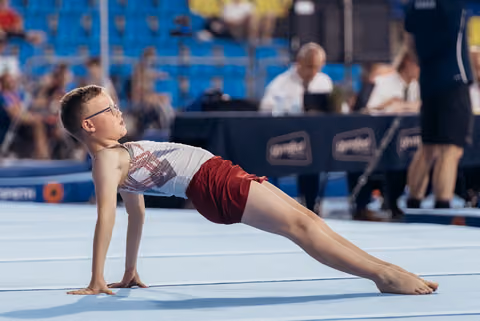 Young boy with glasses performs a back bridge pose on the floor exercise mat, focused and composed during competition.