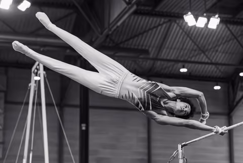 Young gymnast performs a dramatic release move on the high bar, body fully extended in a swan dive position, indoor gym.