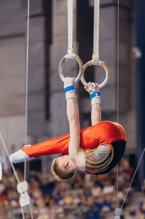 Young male gymnast in red suit performs inverted tuck on still rings, showing intense concentration before a packed crowd.