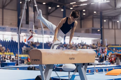 Young male gymnast performs on pommel horse, legs raised high, focused expression, at indoor gymnastics competition.