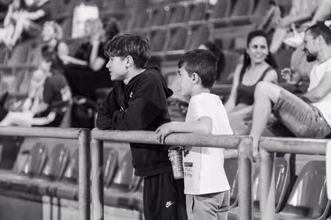 Two boys lean on a railing at a stadium, chatting while watching a sports event, one holding a drink can.