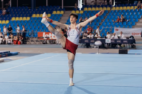 Young male gymnast performs a scale on floor, leg raised high, arms outstretched, focused expression in a blue-seated arena.