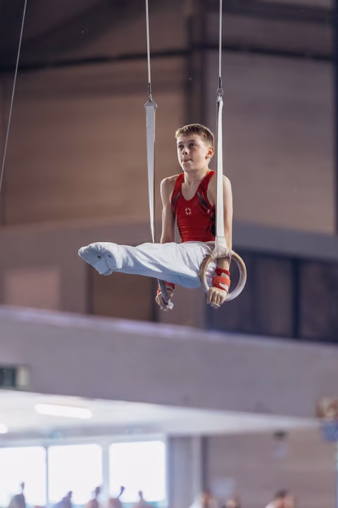 Young male gymnast performs an L-sit on still rings, wearing red leotard, with intense concentration in a gymnasium.