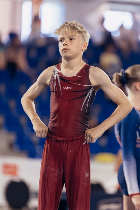 Young male gymnast in dark red Agiva suit stands confidently, hands on hips, gazing intently ahead in a busy sports arena.