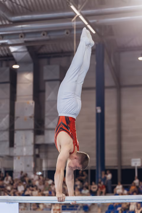 Young male gymnast performs a perfect handstand on parallel bars, legs split, intense concentration visible on his face.
