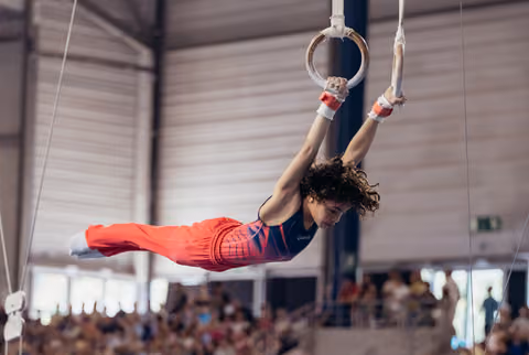 Young male gymnast performs an iron cross on rings, body horizontal, intense focus, in a packed indoor arena.