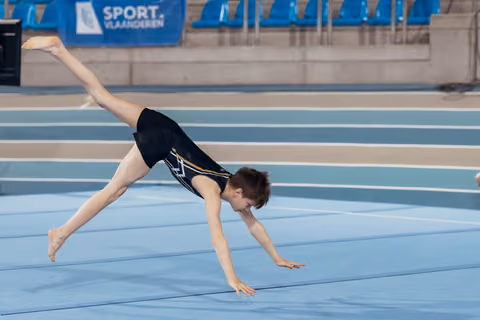 Young gymnast performs a cartwheel on the blue floor mat at a Sport Vlaanderen indoor gymnastics event.