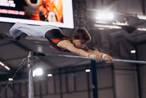 Young male gymnast grips the high bar with intense focus, body horizontal in a dramatic release position indoors.