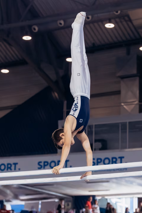 Young male gymnast performs a precise handstand on parallel bars, legs perfectly vertical, in an indoor sports hall.