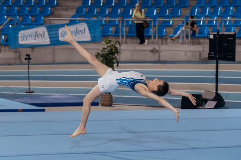 Young gymnast performs a split leap on the floor exercise, body arched gracefully in a white and blue leotard.