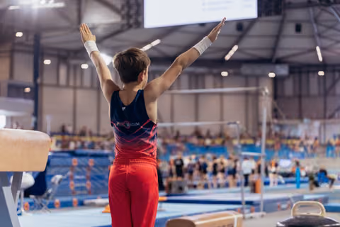 Young male gymnast raises both arms in a triumphant salute inside a large gymnastics arena, wearing red and navy.