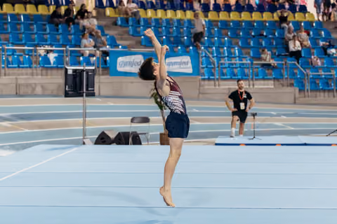 Young male gymnast lands with arms raised triumphantly on the floor exercise mat during a gymnastics meet.