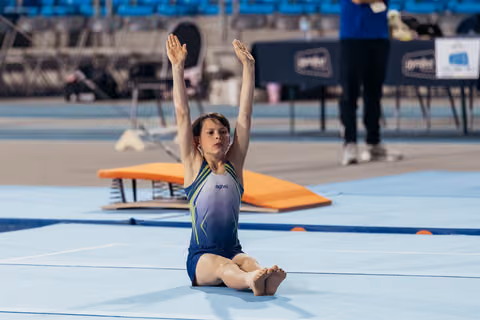 Young gymnast sits on blue floor mat, arms raised high with focused expression, wearing blue agiva leotard.