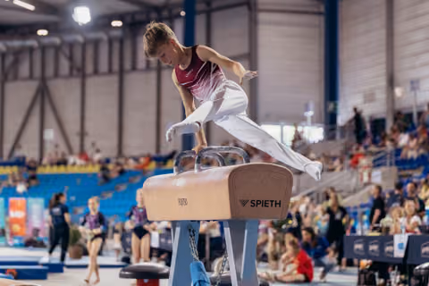Young male gymnast performs on pommel horse, leaning forward with intense focus during an indoor gymnastics meet.