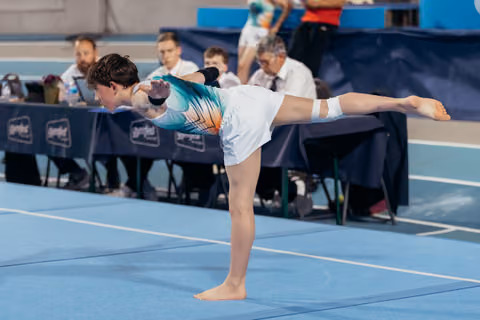 Young gymnast holds a precise arabesque on blue floor, leaning forward with arms extended, focused during competition.