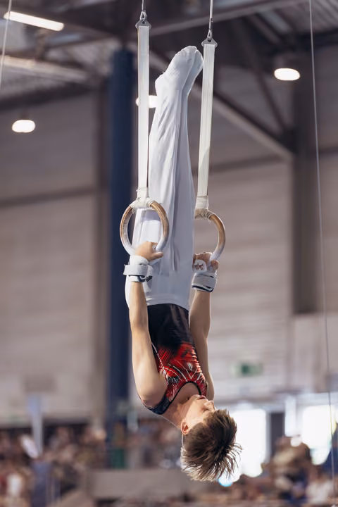 Young male gymnast performs an inverted hang on still rings, body fully extended, in a packed competition gymnasium.