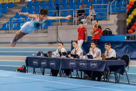Male gymnast soars mid-air in a swan dive above judges at a Gymfed competition, arms outstretched with focus.